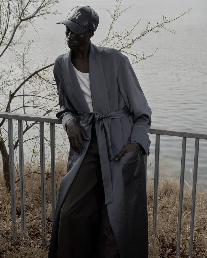Elegant dark-skinned adult wearing a robe and baseball cap, leaning on a railing with lake background. Fashion and lifestyle imagery.