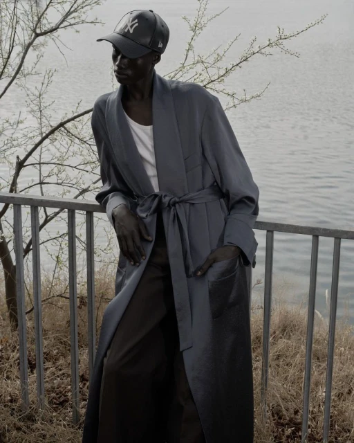 Elegant dark-skinned adult wearing a robe and baseball cap, leaning on a railing with lake background. Fashion and lifestyle imagery.