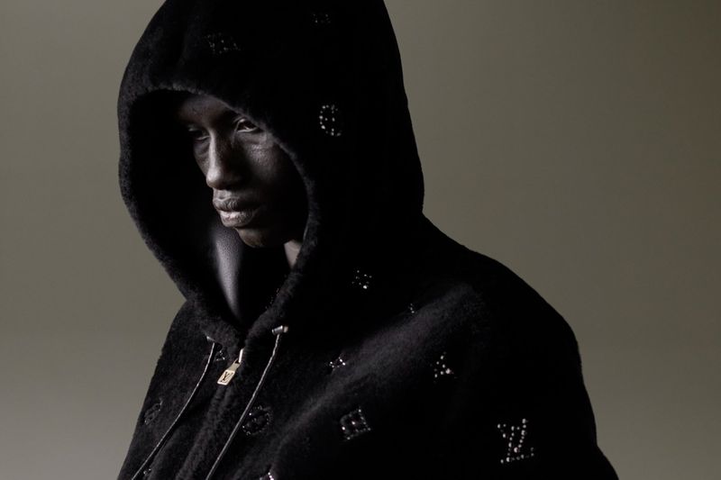 Close-up fashion portrait of an adult female model with dark skin and a dark hoodie with monogram details in a studio setting.