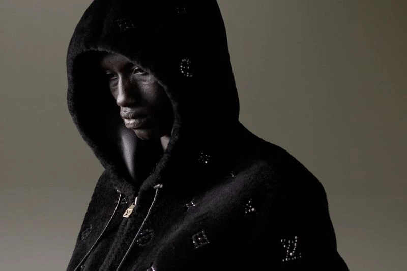 Close-up fashion portrait of an adult female model with dark skin and a dark hoodie with monogram details in a studio setting.