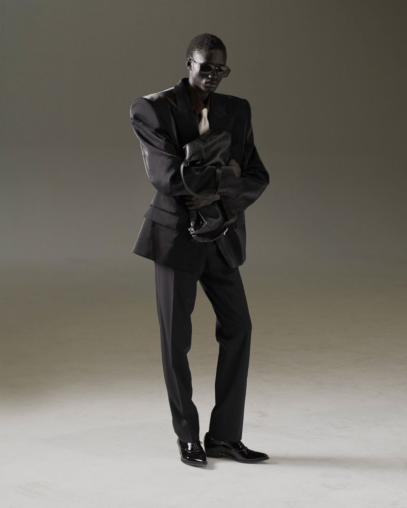 An adult male fashion model in a dark suit poses in a studio, holding a black bag. The low-angle shot emphasizes his height and the suit's structure.