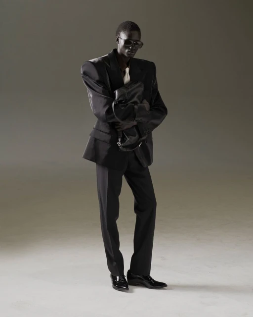 An adult male fashion model in a dark suit poses in a studio, holding a black bag. The low-angle shot emphasizes his height and the suit's structure.