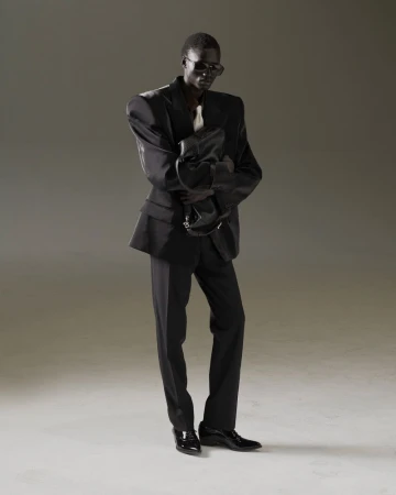 An adult male fashion model in a dark suit poses in a studio, holding a black bag. The low-angle shot emphasizes his height and the suit's structure.