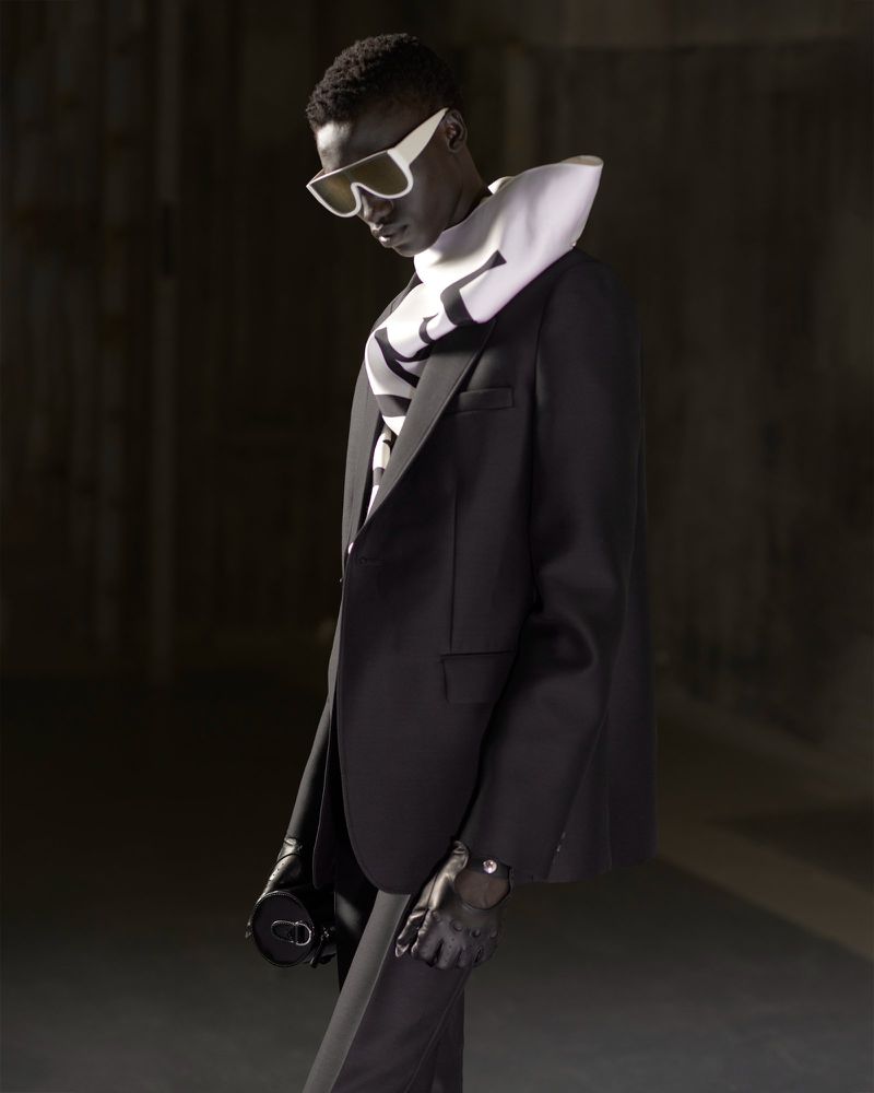 Dark-skinned adult male fashion model in black suit and white-framed sunglasses, against a dark background, shot in studio lighting.