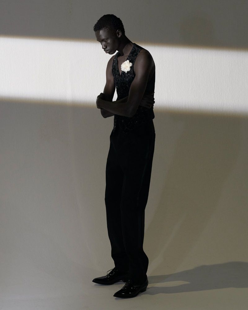 Young adult Black male in formal black attire with a white flower, posing in a photo studio with a neutral background and soft, directed lighting.