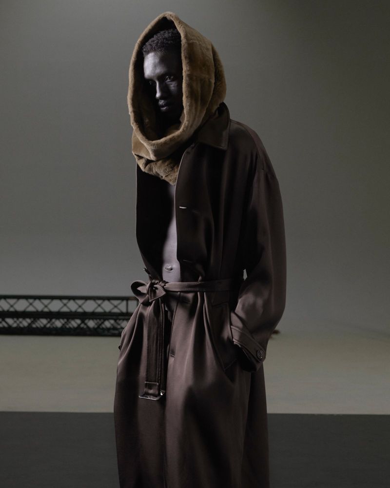 Dark-skinned adult stands in a photo studio wearing a fur-lined hood and a dark brown coat with a sash. Studio lighting.