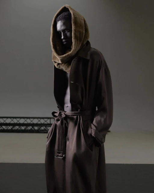Dark-skinned adult stands in a photo studio wearing a fur-lined hood and a dark brown coat with a sash. Studio lighting.