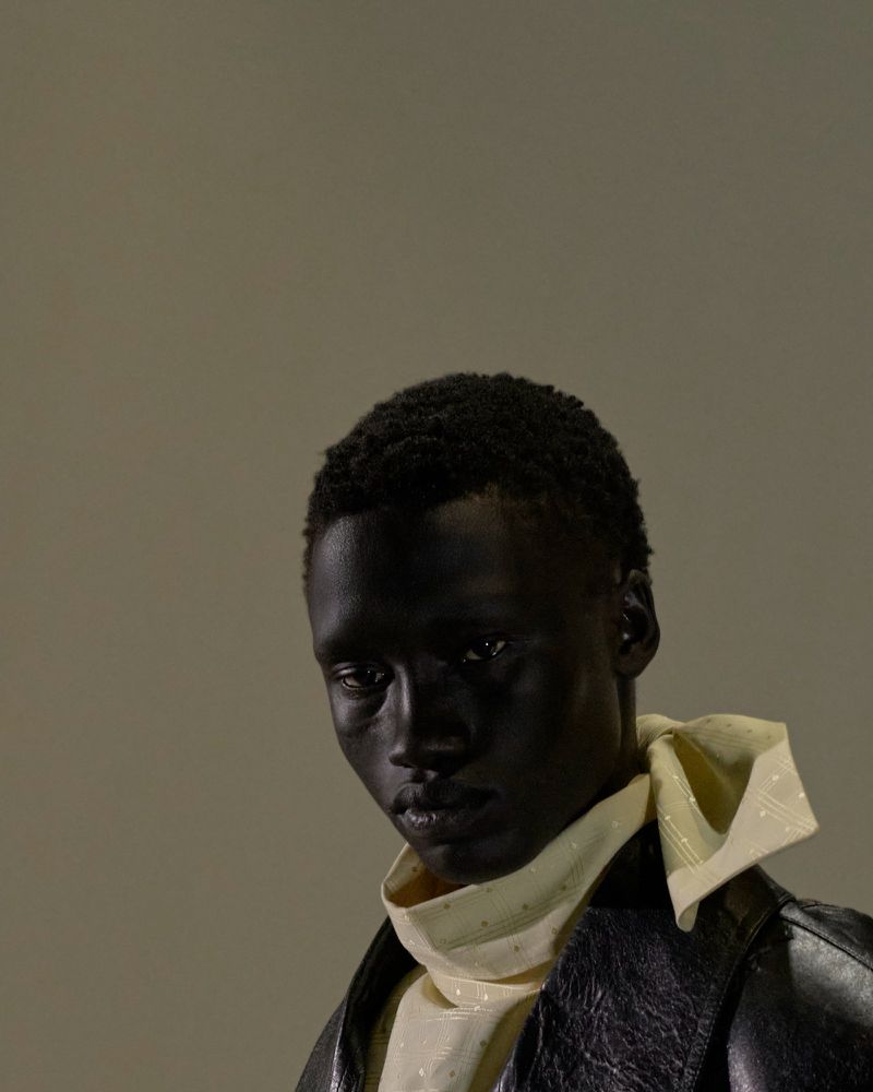 Portrait of a dark-skinned adult male wearing a white collared shirt with a yellow neck scarf and a black leather jacket against grey.
