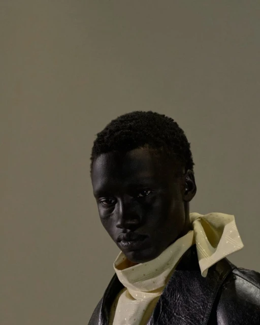 Portrait of a dark-skinned adult male wearing a white collared shirt with a yellow neck scarf and a black leather jacket against grey.