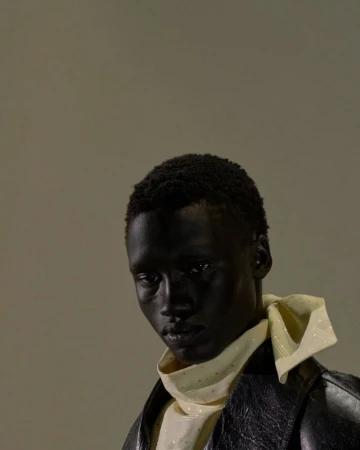 Portrait of a dark-skinned adult male wearing a white collared shirt with a yellow neck scarf and a black leather jacket against grey.