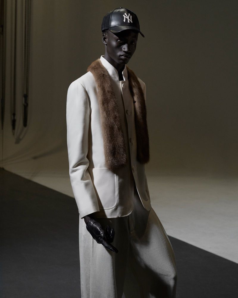 A black male model is posing in a white suit, fur stole and baseball cap against a gradient gray backdrop in a fashion editorial shot.