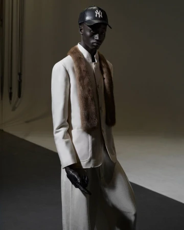 A black male model is posing in a white suit, fur stole and baseball cap against a gradient gray backdrop in a fashion editorial shot.