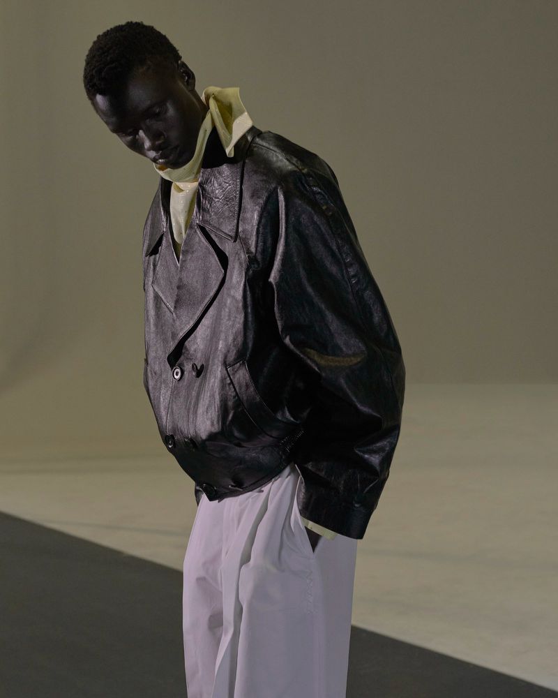Fashion portrait of a black male model in a black leather jacket and white pants standing in a photo studio with neutral lighting.
