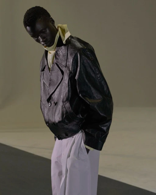 Fashion portrait of a black male model in a black leather jacket and white pants standing in a photo studio with neutral lighting.