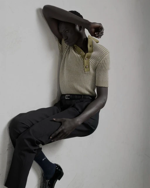 Dark-skinned adult male fashion model poses against a wall wearing a tan collared shirt and black pants with patent leather shoes.