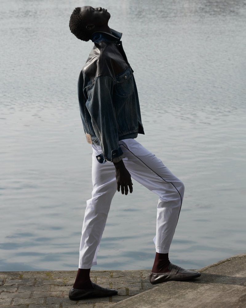 Fashion shot of a male model wearing a denim jacket and white pants posing outdoors by a river, with an upward gaze.