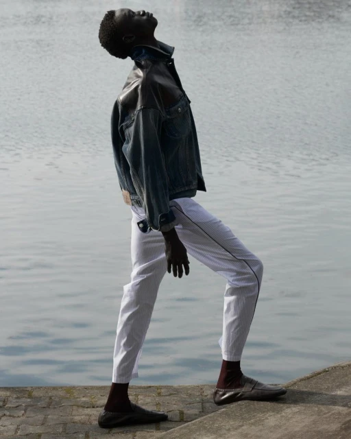 Fashion shot of a male model wearing a denim jacket and white pants posing outdoors by a river, with an upward gaze.