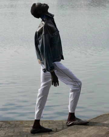 Fashion shot of a male model wearing a denim jacket and white pants posing outdoors by a river, with an upward gaze.