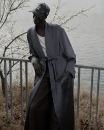 Model in grey belted robe jacket and black baseball cap leaning against metal railing outdoors near water.