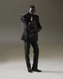 Black man in oversized black suit jacket and trousers holding a dark leather bag against a neutral studio background.