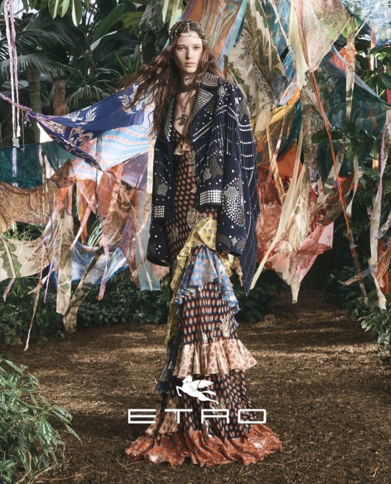 A woman in a patterned coat and dress stands in a wooded area surrounded by draped fabrics, showcasing fashion design.