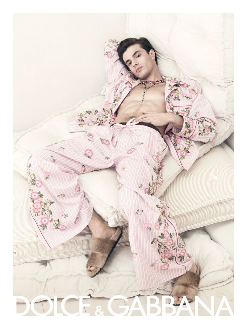Male model in floral pajamas lounged on a stack of white cushions; fashion editorial shot, bedroom setting, warm lighting, designer Dolce & Gabbana.