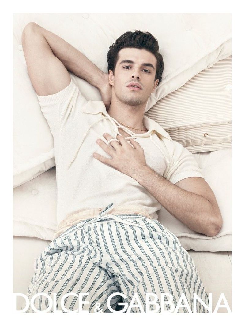 Attractive man lying on white bedding wearing a cream-colored polo-style shirt and striped pyjama pants in a fashion shot.