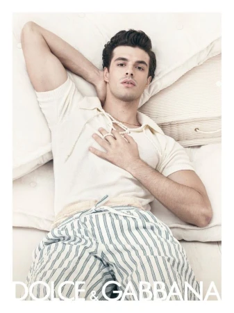 Attractive man lying on white bedding wearing a cream-colored polo-style shirt and striped pyjama pants in a fashion shot.