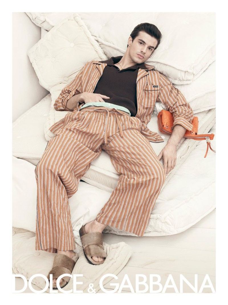 Male fashion model lies on a bed of fluffy pillows wearing striped pajamas, brown slides, and a brown collared shirt in a studio shot.