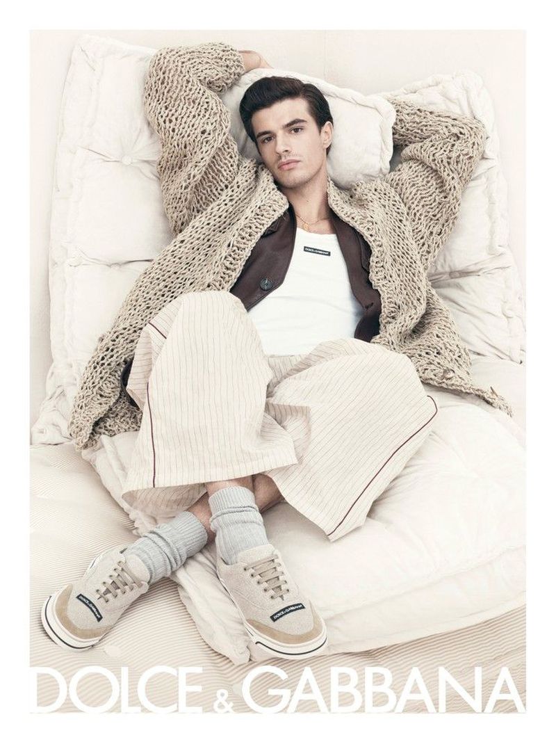 A man lies on a white quilted bed wearing a knit cardigan, brown jacket, white tank top, striped pajamas, socks, and tennis shoes.