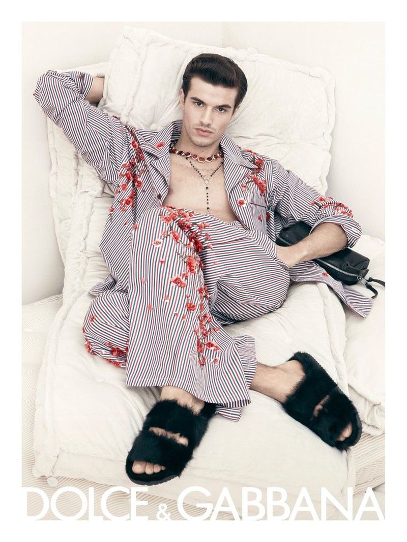 Adult male fashion model lounges on white bedding wearing striped pajamas, furry slides, and jewelry, bathed in soft, natural light.