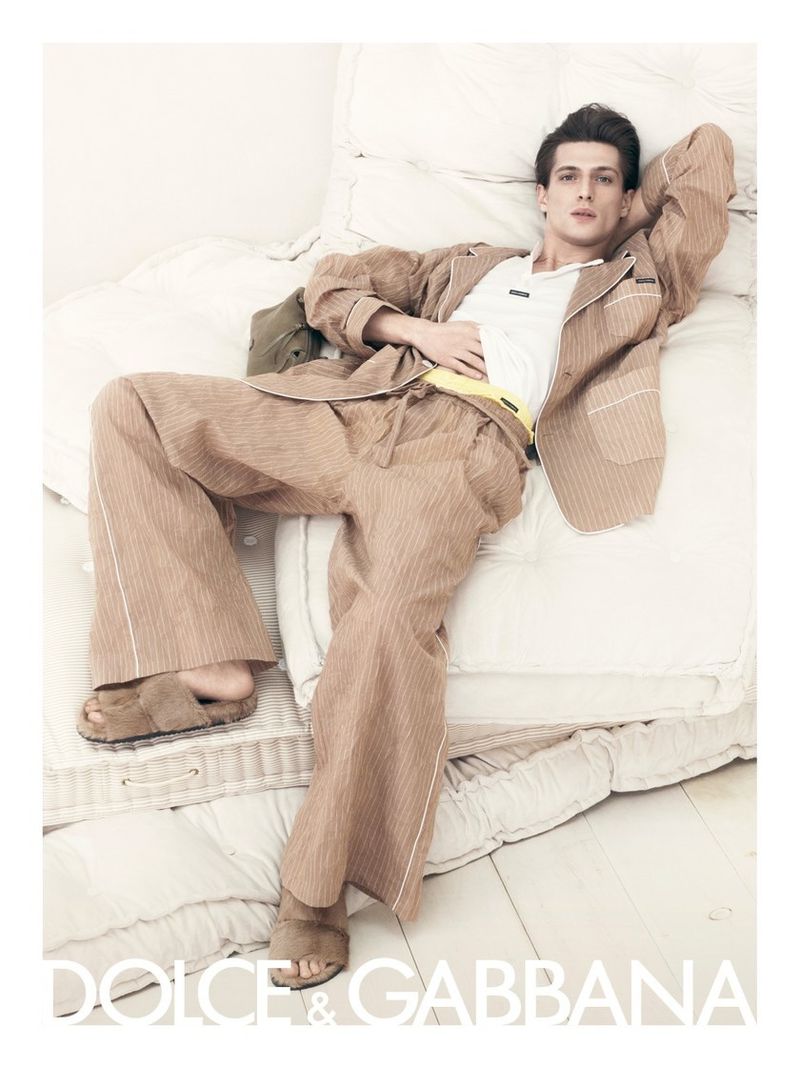 Man poses on cushions wearing a striped pajama-style suit with tan sandals in a fashion editorial ad for Dolce & Gabbana.