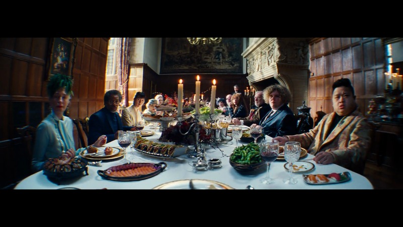 Elegant dinner scene with group of diverse people seated at a long table filled with food and candles in a grand dining hall.