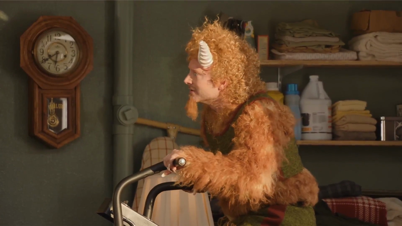 A man in a furry costume with horns stands indoors next to an exercise machine, with a wooden clock and shelves in the background.