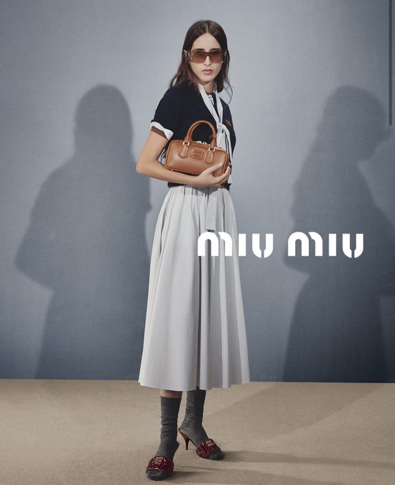 Fashionable woman in a black top, light grey skirt, sunglasses, and maroon heels holds a small brown handbag in a studio shot.