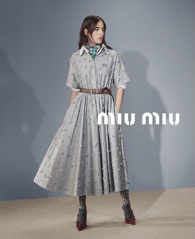 Fashionable woman in a Miu Miu dress with a brown belt, patterned tights, and heeled shoes against a blue background.