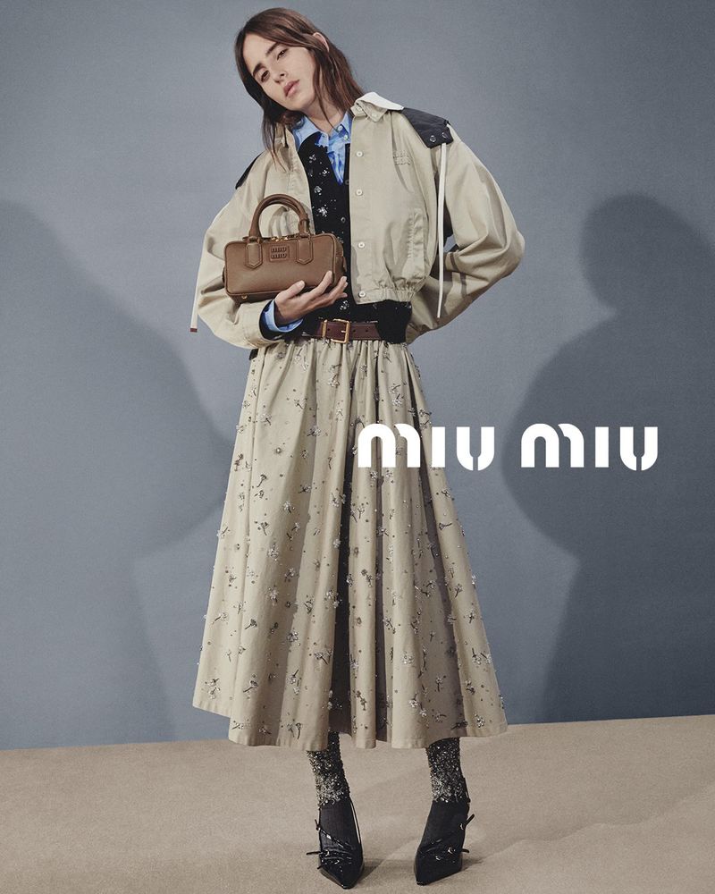 Fashion editorial shot of a woman posing with a Miu Miu handbag, wearing a skirt, stockings, and a jacket against a muted background.