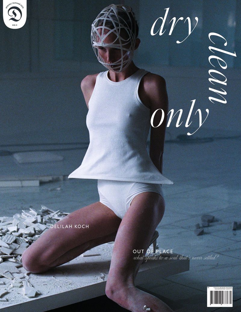 Editorial fashion shot of a woman in a white outfit and geometric face cage kneeling on broken white tiles in an empty room.