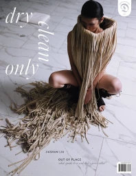 Model kneeling on a white tiled floor wearing a sculptural fringed garment cascading down around her