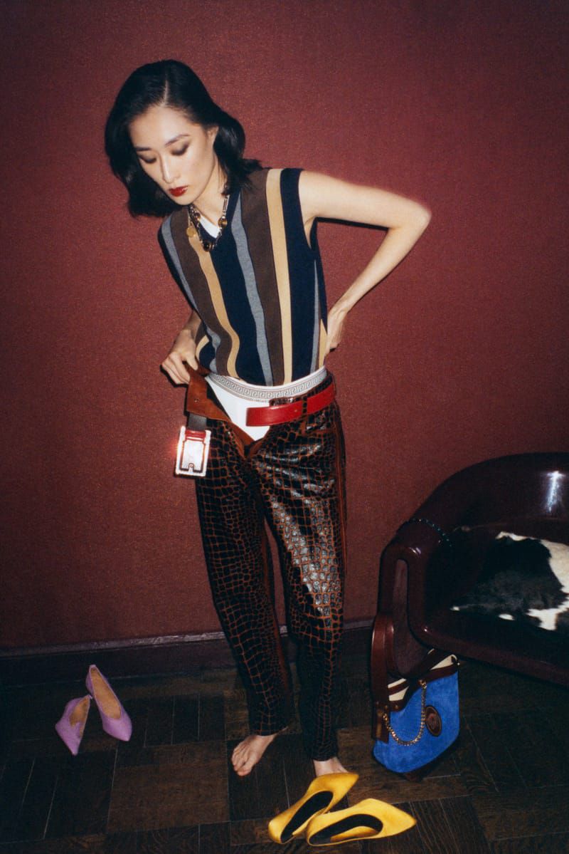 An Asian woman poses in a vintage fashion outfit, wearing a striped vest, patterned pants, and heels, with a red wall backdrop.