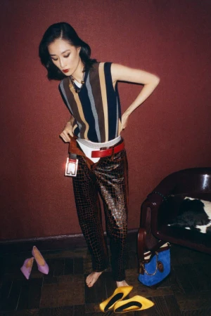 An Asian woman poses in a vintage fashion outfit, wearing a striped vest, patterned pants, and heels, with a red wall backdrop.