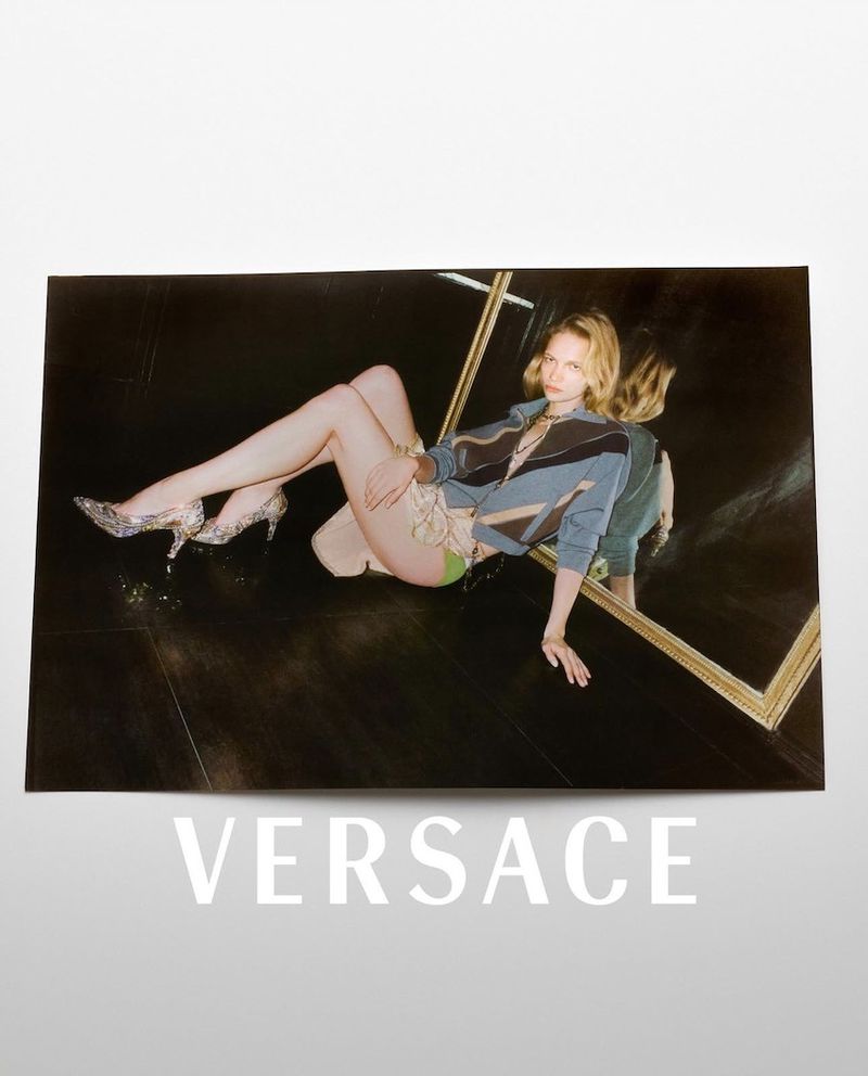 Fashion shot of a blond woman in casual clothing and glitter heels lying on a reflective black floor with a mirror in the background.