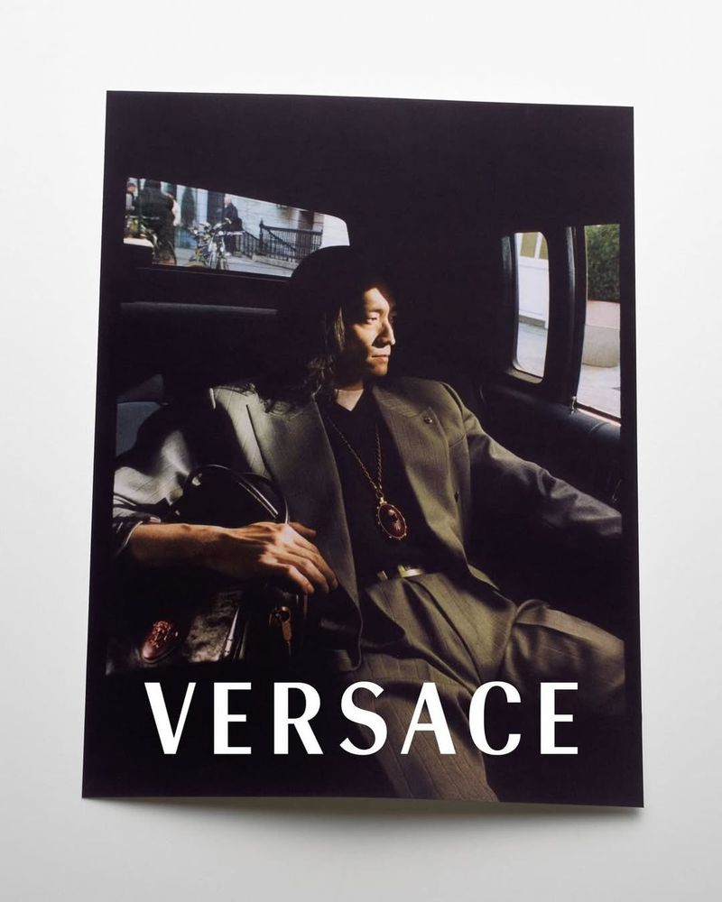 A man wearing a grey suit jacket and a pendant necklace seated inside a vehicle with the brand Versace displayed.