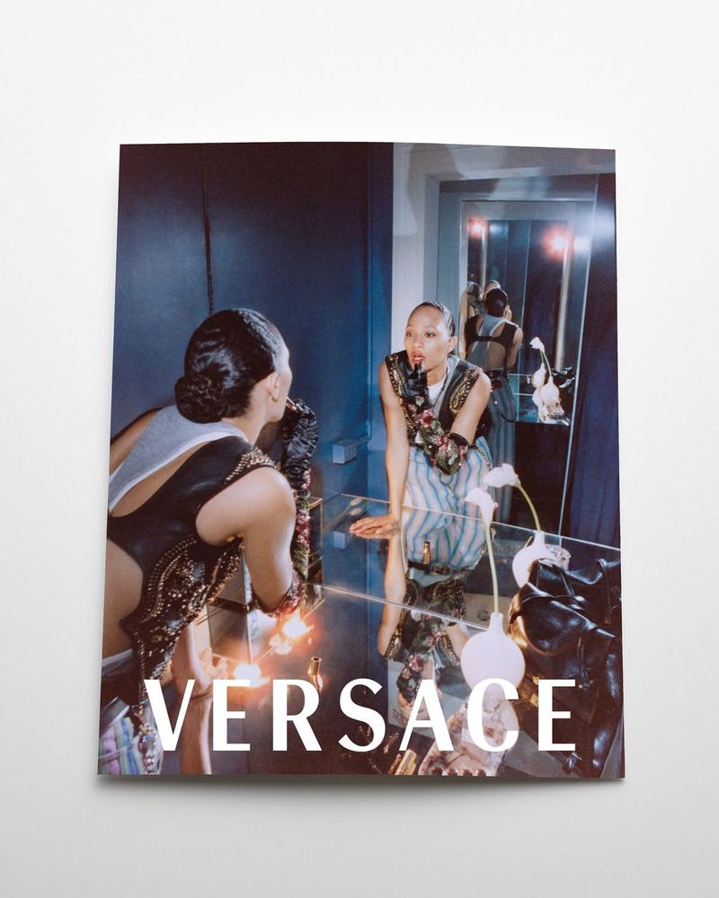 Versace fashion magazine ad: A woman in a decorative bodysuit poses near a mirror, reflecting her image in a blue-toned room.