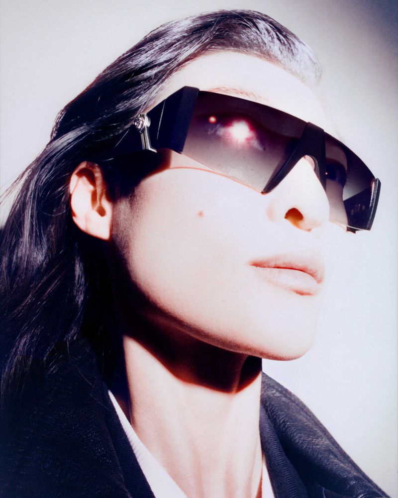 Close-up portrait of a model wearing dark futuristic sunglasses and a black jacket against a plain background