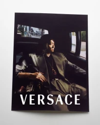 A man wearing a grey suit jacket and a pendant necklace seated inside a vehicle with the brand Versace displayed.