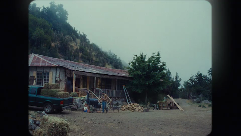 Man tending a barbecue grill near a rustic cabin with a truck, wood pile, and trees under an overcast sky in a rural environment.