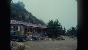 A man stands near a green pickup truck loaded with hay bales beside a rustic house on a rural hillside under an overcast sky.