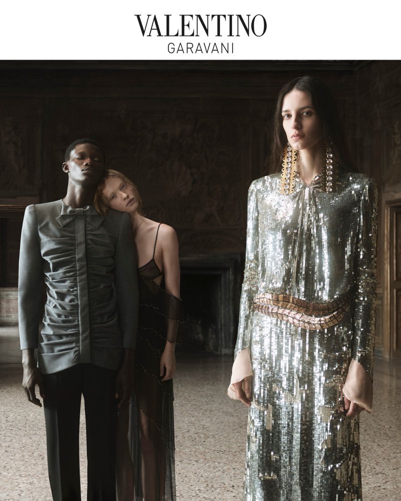 Valentino Garavani fashion advertisement featuring three models wearing elegant gowns and suits in a dimly lit interior setting.