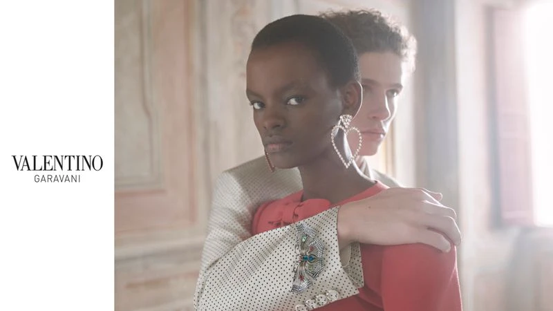Fashion shot of an African American woman in a red dress, wearing large heart-shaped earrings and a brooch, with a male figure in the background.